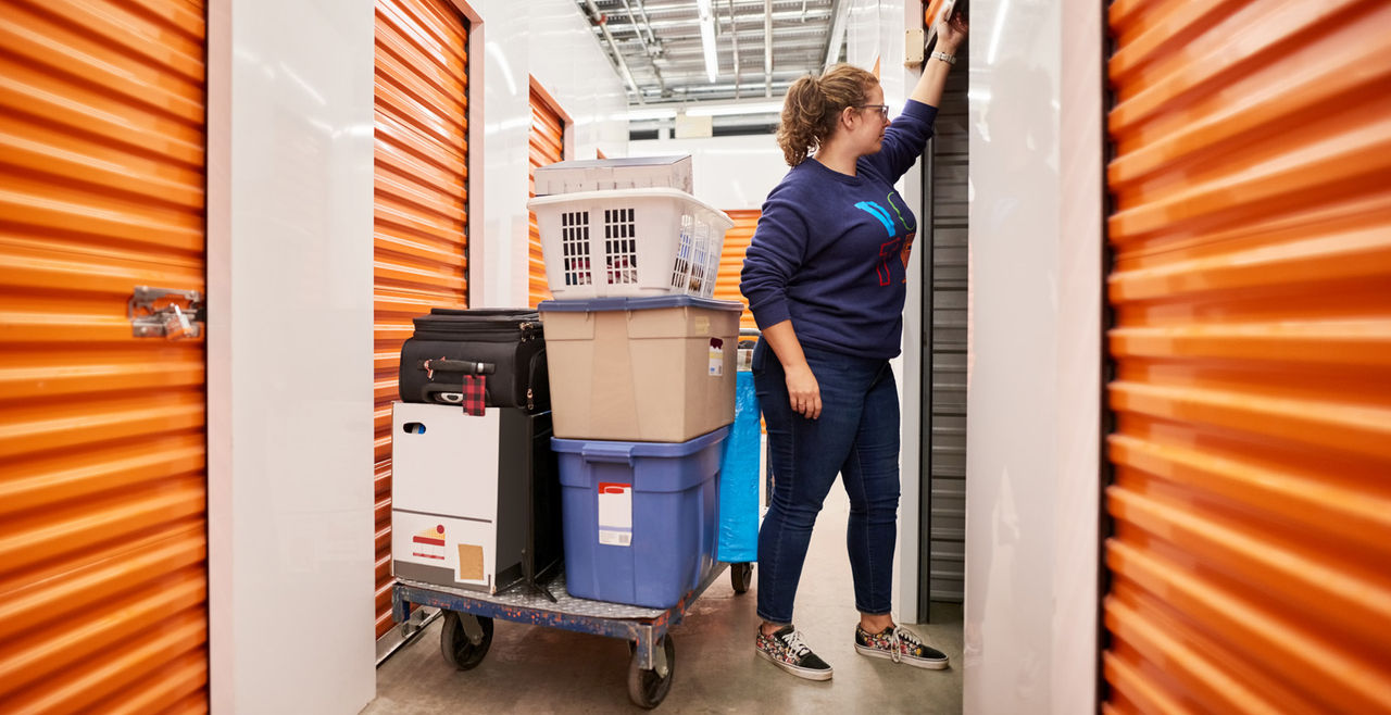 Woman opening the shutter of a self storage unit with a trolley full of goods and items in the aisle of self-storage building